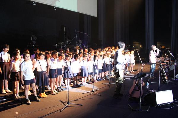 神戸国際会館のホールで「ちめいど」の二人が歌い、後ろには制服を着た子供達が一緒に歌っている写真